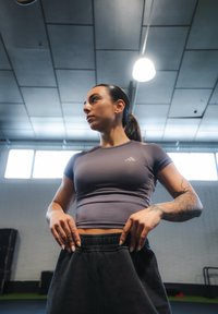 Grå atletisk topp med korte ermer, med en diskret logo, kombinert med svarte joggebukser med snøring. Bakgrunnen viser treningsutstyr.