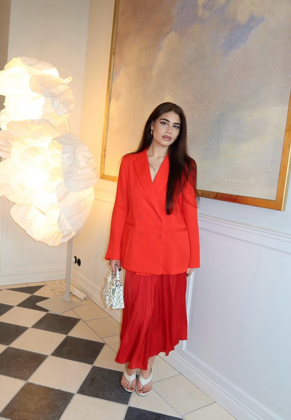 Femme aux longs cheveux bruns, portant un blazer et une jupe rouge vif, tenant un petit sac blanc et debout à l'intérieur près d'une lampe lumineuse et d'un tableau.