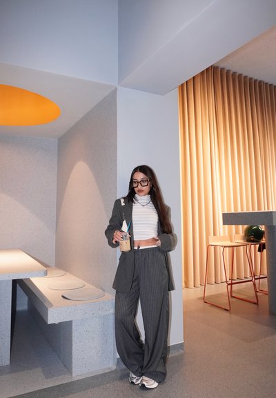Blazer gris a rayas y pantalones de pierna ancha, combinados con un top corto de canalé blanco. Sosteniendo una bebida, en un interior minimalista y moderno.