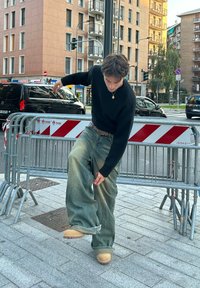 Afslappede, falmede blå jeans, sort sweater og brune klipklapper. Person justerer buksebenet nær byggehegn på en pavébelagt fortov.
