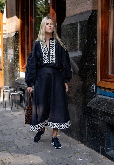 Vestido de lino negro con mangas largas y abullonadas, que presenta detalles bordados en blanco y una falda acampanada. Combinado con zapatillas negras y un bolso de mimbre.