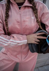 Rozā satīna sporta jaka ar baltām svītrām, augstu apkakli un rāvējslēdzēju. Persona tur austiņas vienā rokā un brūnu priekšmetu otrā.