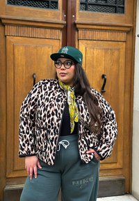 Leopardenmuster Steppjacke mit schwarzen Akzenten, kombiniert mit einem schwarzen Oberteil, grünen Jogginghosen und einer grünen Mütze. Mit einem gemusterten Schal.