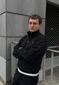 Sweat-shirt noir à fermeture éclair avec logo Nike, porté par-dessus un t-shirt blanc. Environnement urbain gris avec une clôture métallique et un fond en béton texturé.