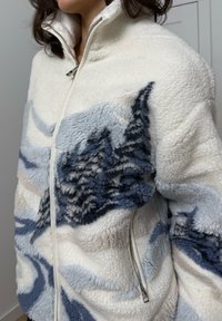 Fleecejakke med en hvid base og blå abstrakte mønstre. Den har en høj krave, lynlåslukning og tekstureret materiale.