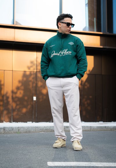 Sudadera de forro polar verde con el logo de "Quiet Place", cuello con cremallera; combinada con pantalones beige claros y zapatos beige, de pie contra un fondo moderno.