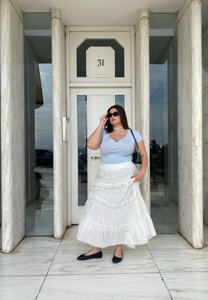 Top de manga corta azul claro con escote en V, falda blanca de volantes con encaje, zapatos planos negros, usando gafas de sol, de pie frente a un edificio.