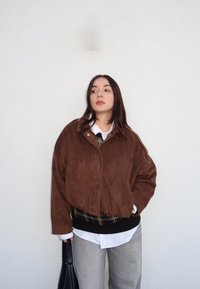 Veste en daim marron à coupe décontractée, fermeture éclair et col ; portée par-dessus une chemise blanche et une ceinture à carreaux noirs ; pantalon gris.