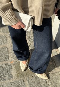 Bézs bordázott pulóver, sötétkék bőszárú farmernadrág, fehér borítéktáska és krémszínű, ezüst szegecses hegyes orrú magassarkú cipő. Texturált és rétegezett megjelenés.