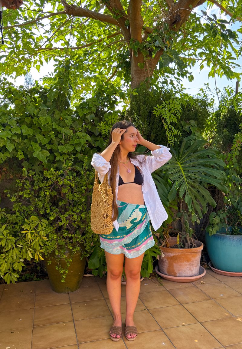 Femme debout sur une terrasse carrelée, ajustant ses cheveux, portant un haut de bikini noir, une chemise blanche ouverte, un sarong à motifs, des sandales, avec un grand sac tressé et un fond de plantes vertes.