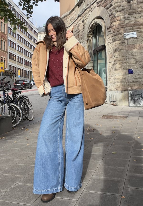 Vrouw in een tan shearling jas, bordeauxrood sweater, wijde blauwe jeans en bruine laarzen die op een stadstoegang loopt, dichtbij fietsen en een stenen gebouw.