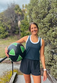 Top sportivo nero con dettagli bianchi, abbinato a pantaloni corti neri. Tiene in mano una grande palla nera con dettagli verdi e bianchi.