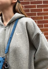 Sweat à capuche zippé gris avec un logo Nike blanc sur la poitrine, accompagné d'un sac à bandoulière bleu avec des ferrures argentées. Tissu texturé.