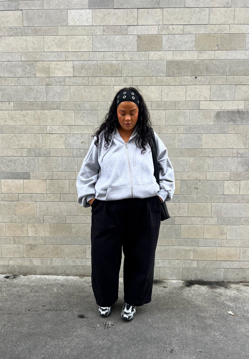 Sweatshirt cinza com fecho éclair e design oversized, combinado com calças largas pretas e sapatos com padrão preto e branco. Mochila texturizada preta pendurada no ombro.