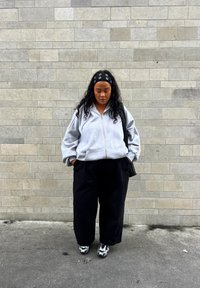 Sweatshirt cinza com fecho éclair e design oversized, combinado com calças largas pretas e sapatos com padrão preto e branco. Mochila texturizada preta pendurada no ombro.