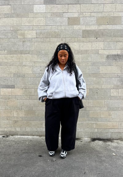 Sudadera gris con cremallera y diseño oversized, combinada con pantalones holgados negros y zapatos de patrón en blanco y negro. Bolsa texturizada negra colgada del hombro.