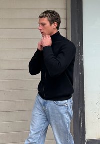 Άνδρας διορθώνει το γιακά του μαύρου πουλόβερ του, στέκεται πλευρικά σε μπεζ τοίχο και κάσα πόρτας, φορώντας ανοιχτόχρωμο τζιν.