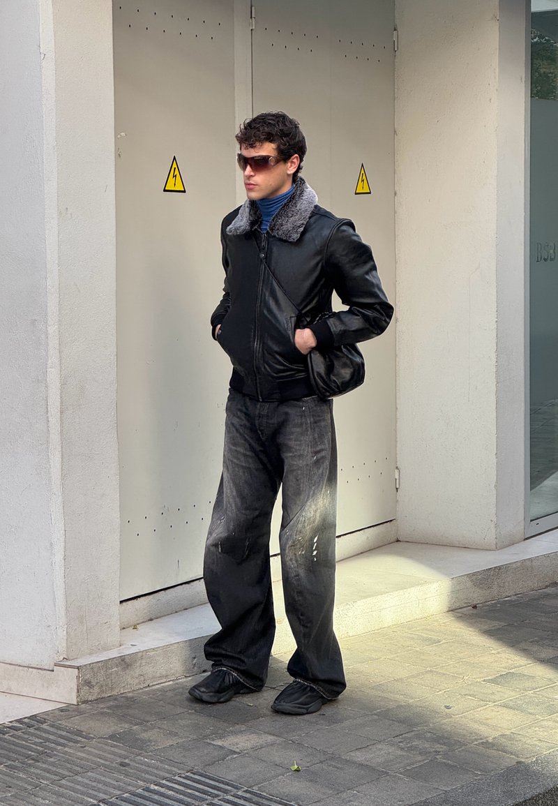 Νεαρός άνδρας με γυαλιά ηλίου, μαύρο δερμάτινο μπουφάν με γούνινο γιακά και φαρδιά μαύρα τζιν, στέκεται με τα χέρια στις τσέπες κοντά σε κλειστή ηλεκτρική καμπίνα.
