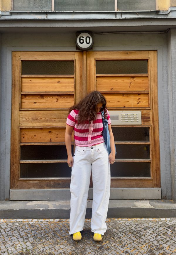Mujer con pantalones blancos y camisa de rayas rosas de pie sobre adoquines frente a puertas dobles de madera numeradas 60, mirando hacia abajo.