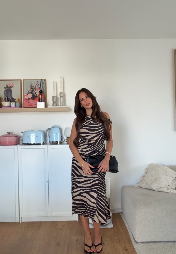 Mujer con cabello largo y oscuro, con un vestido de estampado de cebra, sosteniendo un bolso de mano negro, de pie en una cocina moderna con electrodomésticos azules y suelos de madera clara.