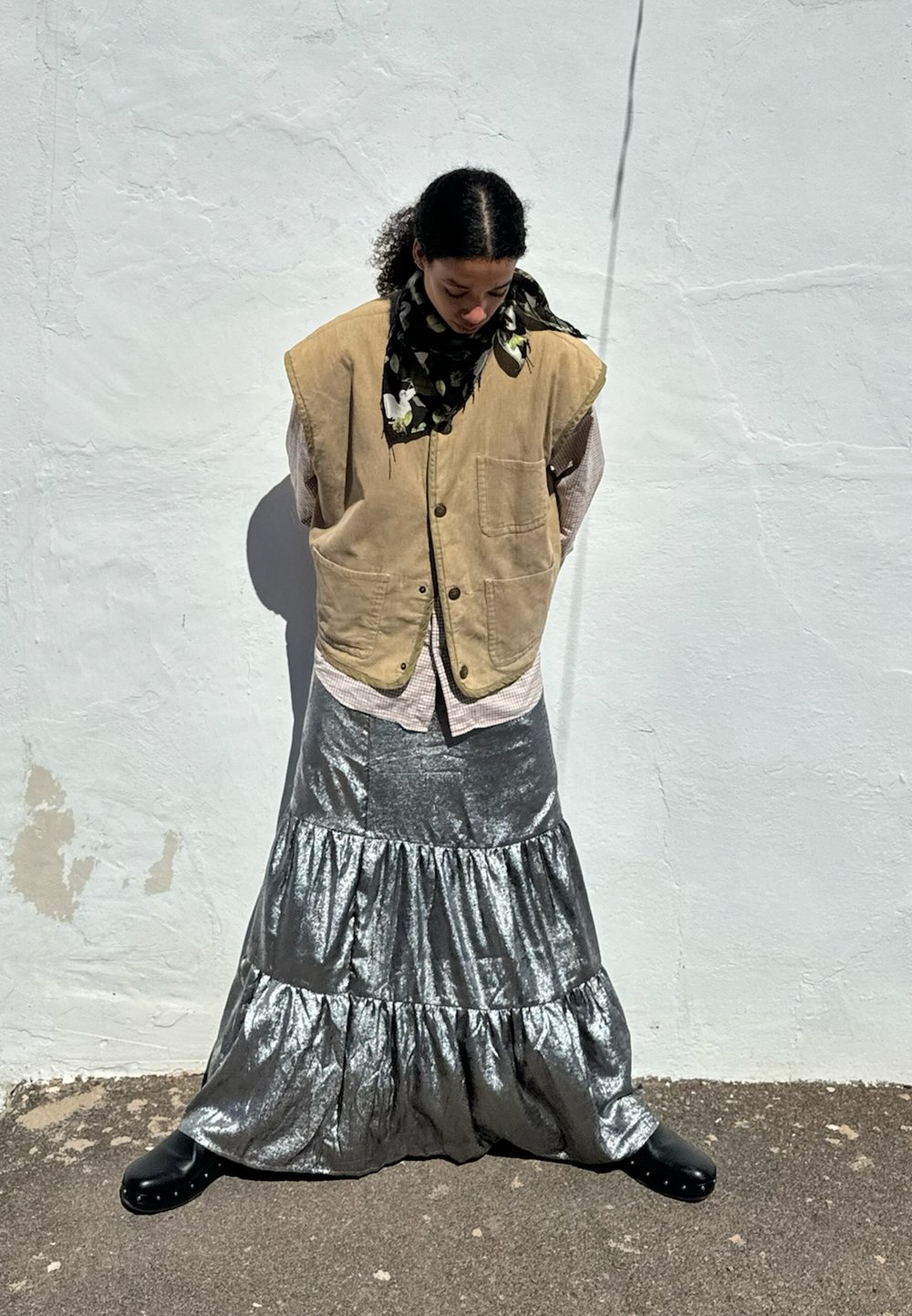 Beige kordflorsväst över en ljusrutigt skjorta, med en metallisk silverfärgad volangkjol och svarta träskor, kompletterat med en blommig scarf.