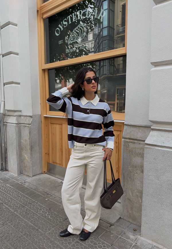 Donna che indossa un maglione a righe bianche e nere, pantaloni color crema, scarpe nere e occhiali da sole, in piedi davanti a una porta di legno con il cartello "Oyster Bar".