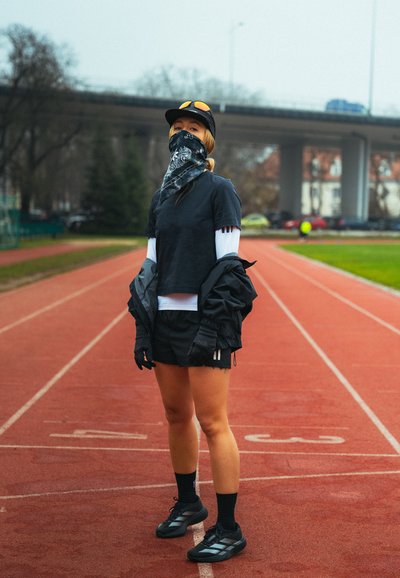Corredor con ropa deportiva negra, guantes y bufanda en la cara, de pie en una pista al aire libre con un paso elevado y árboles al fondo.