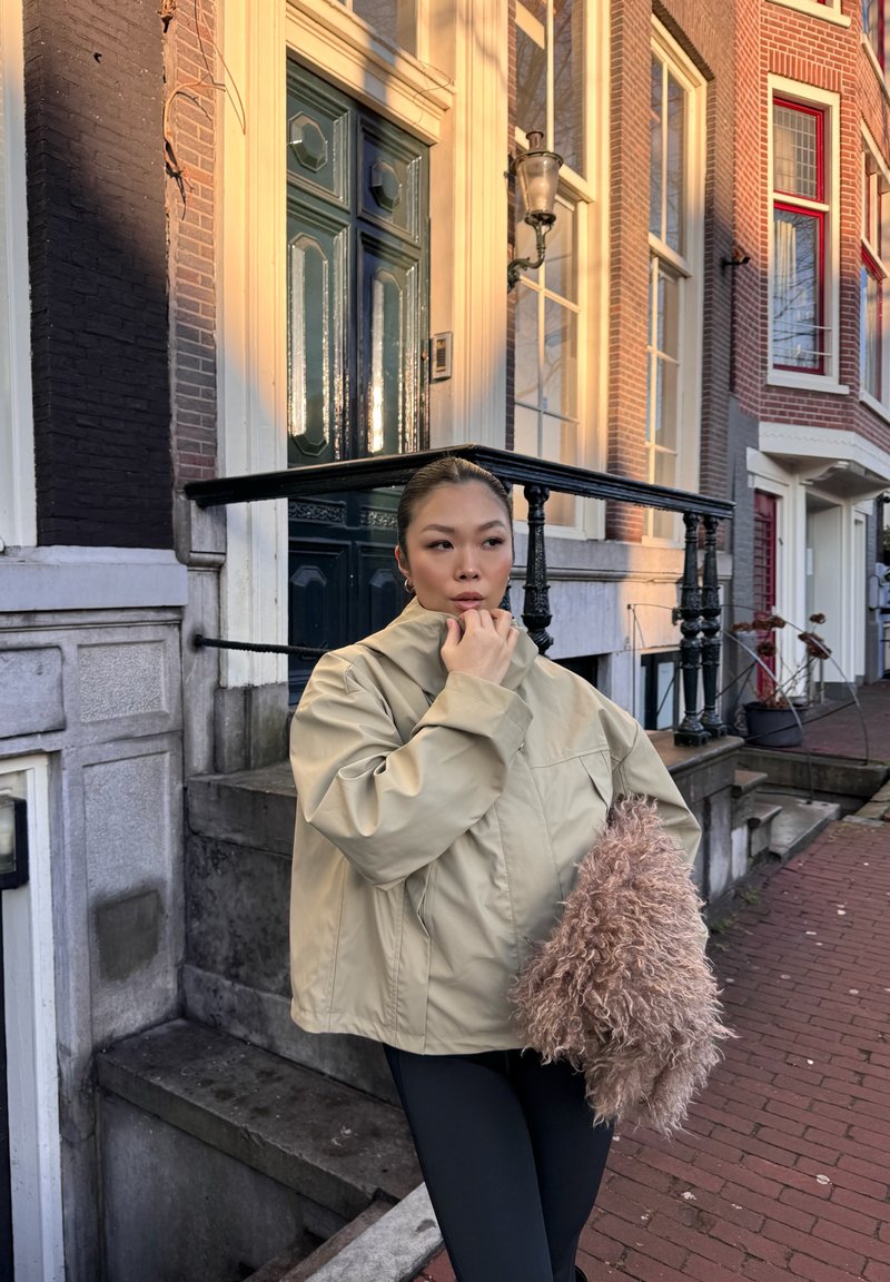 Donna con giacca beige e pantaloni neri in piedi su un marciapiede di mattoni, che tiene una borsa marrone pelosa, accanto a gradini e porta verde scuro di un edificio di mattoni.