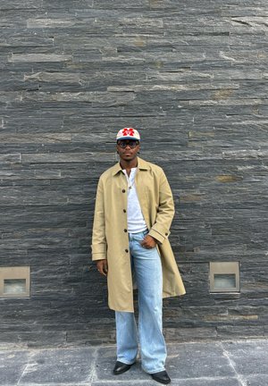 Hombre con abrigo beige, camisa blanca, vaqueros azul claro, zapatos negros, gafas y gorra blanca, de pie frente a una pared de piedra gris oscuro.
