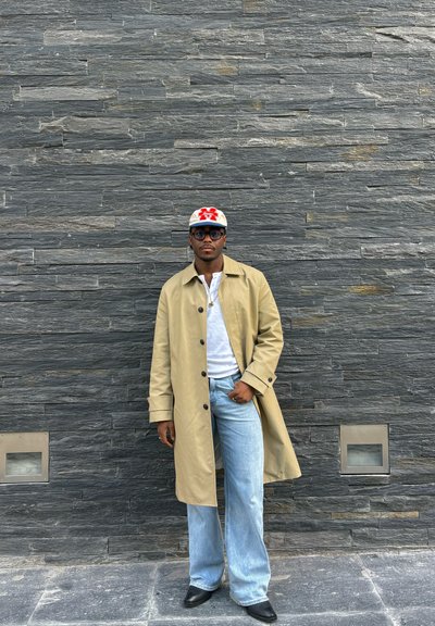 Hombre con abrigo beige, camisa blanca, vaqueros azul claro, zapatos negros, gafas y gorra blanca, de pie frente a una pared de piedra gris oscuro.