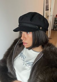 Personne aux cheveux courts et foncés portant une casquette noire, un pull blanc avec un motif gris, et un manteau en fourrure brun foncé, regardant sur le côté à l'intérieur.