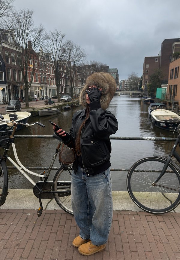 Personne portant une capuche en fourrure et une veste en cuir se tient sur un pont de canal, tenant un téléphone, avec des vélos et des bateaux le long de l'eau dans un cadre urbain nuageux.