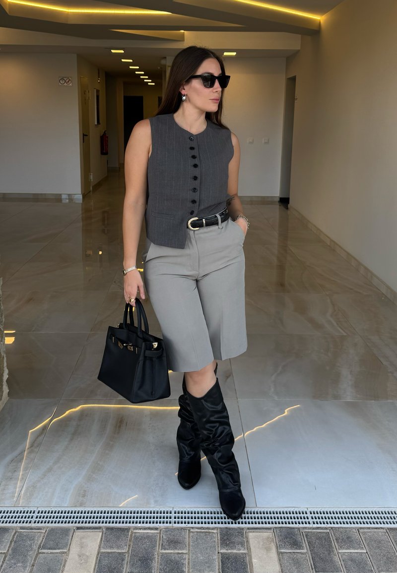 Gilet grigio a righe con bottoni neri, pantaloni beige, stivali neri al ginocchio e una borsa nera con texture. Ambiente interno con illuminazione moderna.