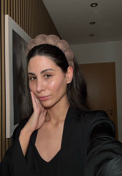 Diadema acolchada marrón, textura suave, usada con una bata negra. El fondo presenta paredes de colores claros y patrones a rayas.