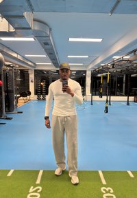 Homme en chemise blanche à manches longues, pantalon de survêtement gris et casquette prenant un selfie devant un miroir dans une salle de sport intérieure avec du matériel de fitness.