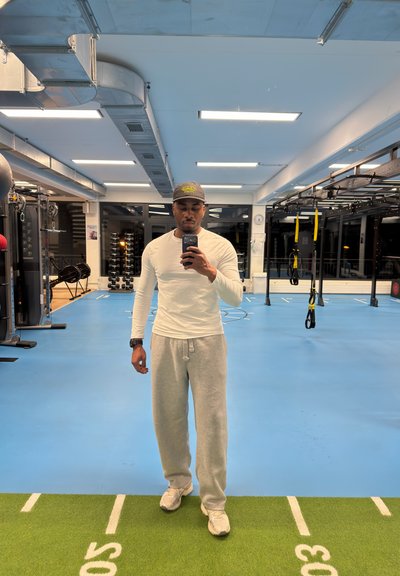 Hombre con camisa blanca de manga larga, pantalones deportivos grises y gorra tomando una selfie frente al espejo en un gimnasio interior con equipo de fitness.
