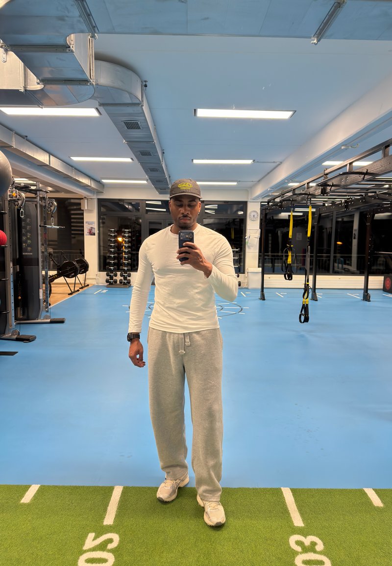 Homme portant une chemise blanche à manches longues, un pantalon de survêtement gris et une casquette, prenant un selfie dans un miroir dans une salle de sport intérieure avec des équipements de fitness.