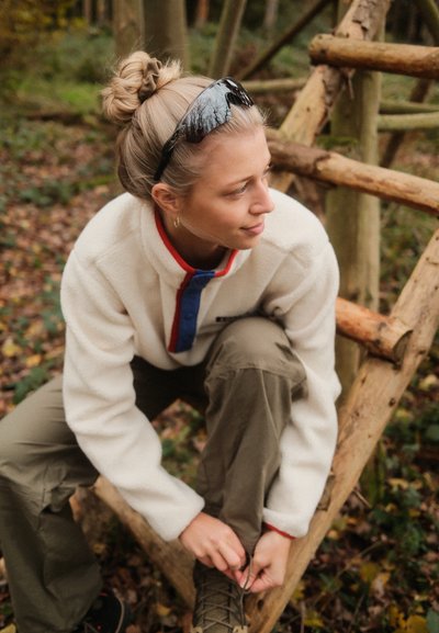 Chaqueta de forro polar color crema con un cuello con cremallera rojo y azul; pantalones verde oliva; botas de senderismo marrones atadas; gafas de sol negras; entorno natural.