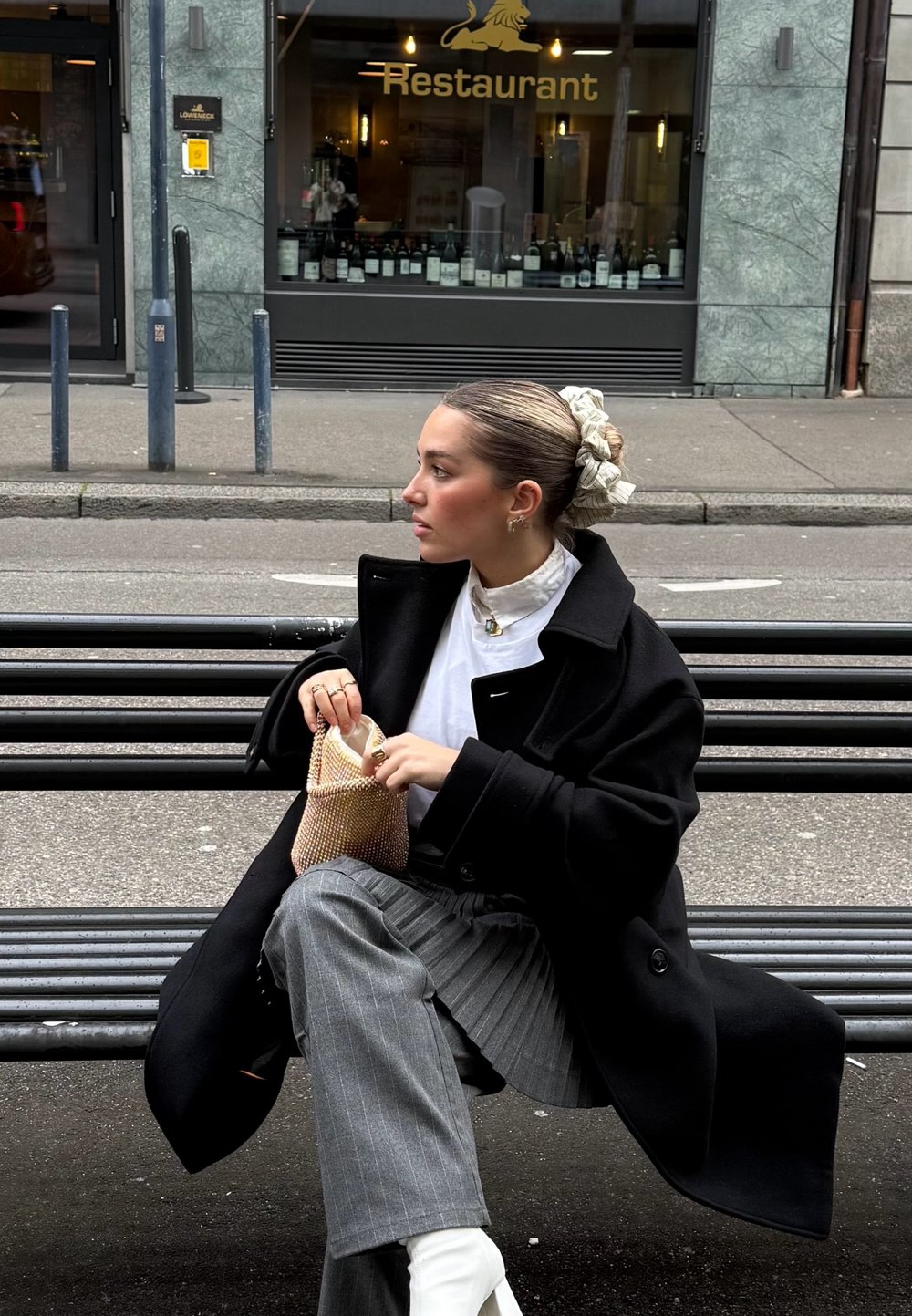 Manteau noir surdimensionné, blouse blanche, pantalon gris à fines rayures, et bottines blanches. Tenant un petit sac en filet, cheveux attachés avec un scrunchie à motif.
