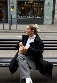 Manteau noir surdimensionné, blouse blanche, pantalon gris à fines rayures, et bottines blanches. Tenant un petit sac en filet, cheveux attachés avec un scrunchie à motif.