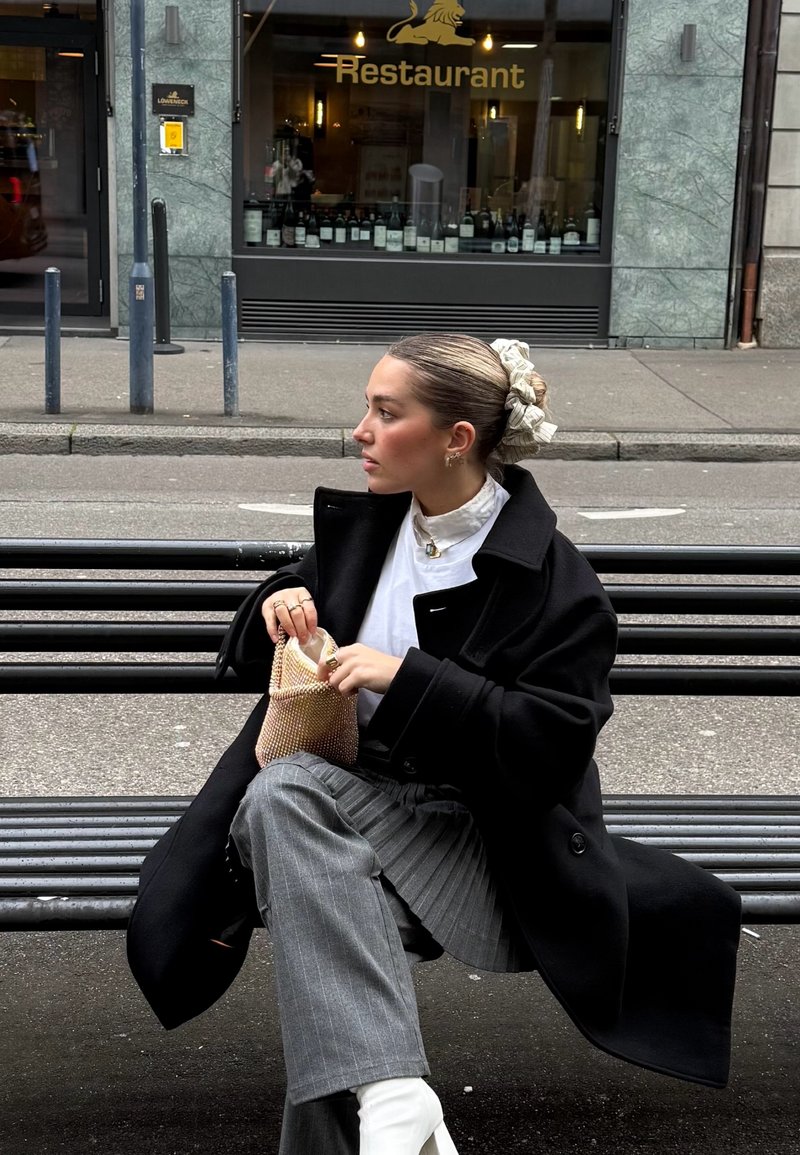 Manteau noir surdimensionné, blouse blanche, pantalon gris à fines rayures, et bottines blanches. Tenant un petit sac en filet, cheveux attachés avec un scrunchie à motif.