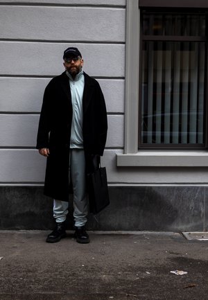 Abrigo largo negro sobre un chándal azul claro, botas negras, llevando una bolsa negra; de pie contra una pared blanca texturizada con una ventana.