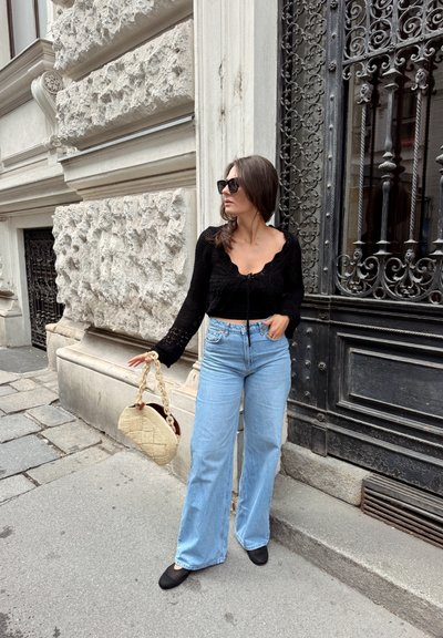 Top negro de tejido de punto con cuello en forma de ondas, vaqueros acampanados azul claro, zapatos planos negros y bolso beige con estampado y asa de cadena.