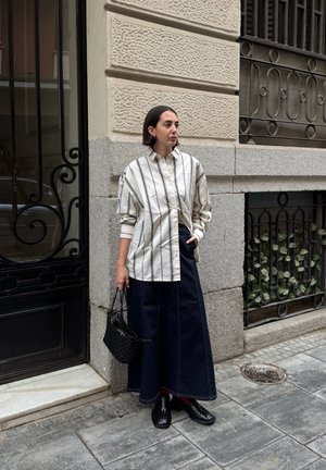 Camisa de botones de gran tamaño a rayas en beige claro y gris; falda de mezclilla oscura; bolso de mano tejido negro; zapatos negros; de pie contra una pared texturizada.