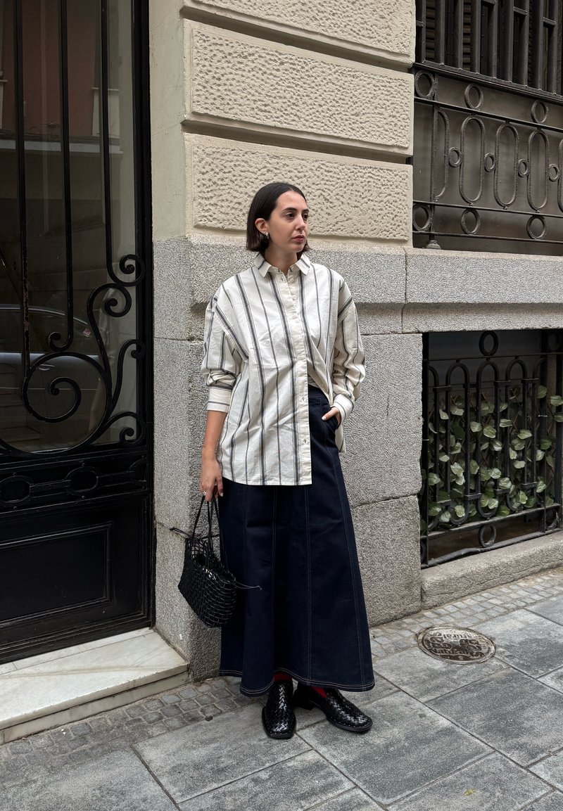 Camisa de botones de gran tamaño a rayas en beige claro y gris; falda de mezclilla oscura; bolso de mano tejido negro; zapatos negros; de pie contra una pared texturizada.