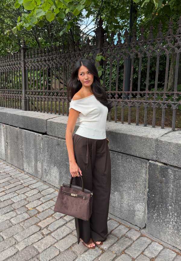 Mujer con cabello largo y oscuro, lleva una blusa blanca sin hombros y pantalones negros, está de pie en un camino de adoquines sosteniendo un bolso marrón junto a una pared de piedra y una valla de hierro.