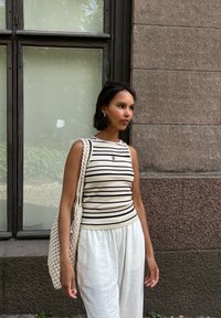 Top de tirantes beige a rayas con un diseño ajustado, combinado con pantalones blancos holgados y un bolso de hombro de mimbre, todo frente a una pared texturizada.