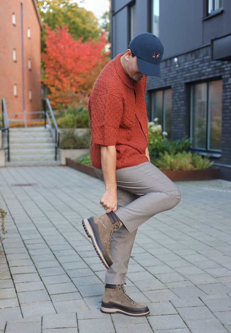 Brown suede hiking boots with metal eyelets, contrasting white sole, and a reinforced heel. Worn with light grey tailored trousers.