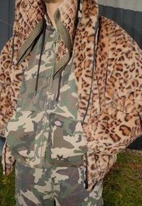 Jakna iz umetnega krzna z leopardjim vzorcem in kapuco, kombinirana s kombinezonom v kamuflažnem vzorcu, ki ima sprednji žep in zavezovalne vrvice za prilagoditev.