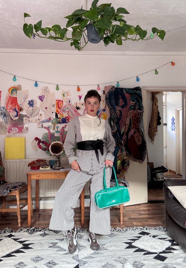 Persona in piedi con abbigliamento a righe e cintura nera in vita che tiene una borsa turchese in una stanza colorata con decorazioni murali, pianta sospesa e mobili in legno.
